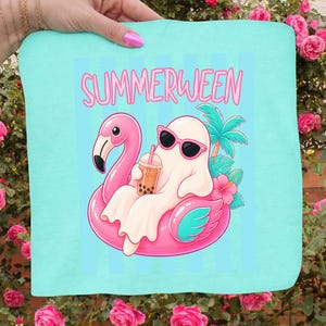 K&ouml;nnte beinhalten: Hellblaues Strandtuch mit dem Wort "SUMMERWEEN" in Rosa. Das Tuch zeigt einen Cartoon-Geist mit Sonnenbrille, der auf einem rosa Flamingo-Schwimmer treibt und Bubble Tea h&auml;lt. Palmen und Hibiskusbl&uuml;ten sind ebenfalls abgebildet.