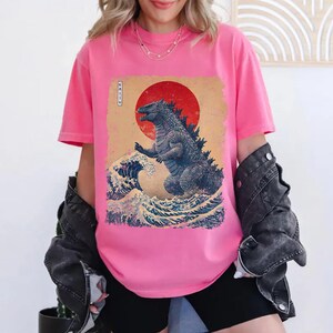 May include: A bright pink t-shirt with a graphic print of Godzilla in a Japanese art style. The design includes a red sun, ocean waves, and the word "KAIJU" in Japanese characters. The shirt is a vibrant pink colour.