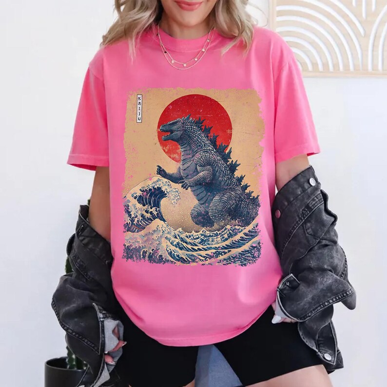 May include: A bright pink t-shirt with a graphic print of Godzilla in a Japanese art style. The design includes a red sun, ocean waves, and the word "KAIJU" in Japanese characters. The shirt is a vibrant pink colour.