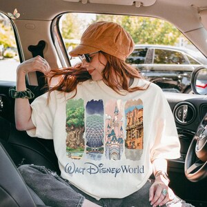 May include: Cream-colored sweatshirt featuring four vertical panels with Disney World imagery: a tree, Spaceship Earth, a castle, and the Hollywood Tower Hotel. The words "Walt Disney World" are printed below the panels.