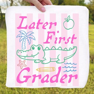 May include: White square towel with pink and white striped background. The towel features the text "Later First Grader" in pink, along with a smiling green alligator, a palm tree, an apple, a starfish, a pencil, and blue waves.