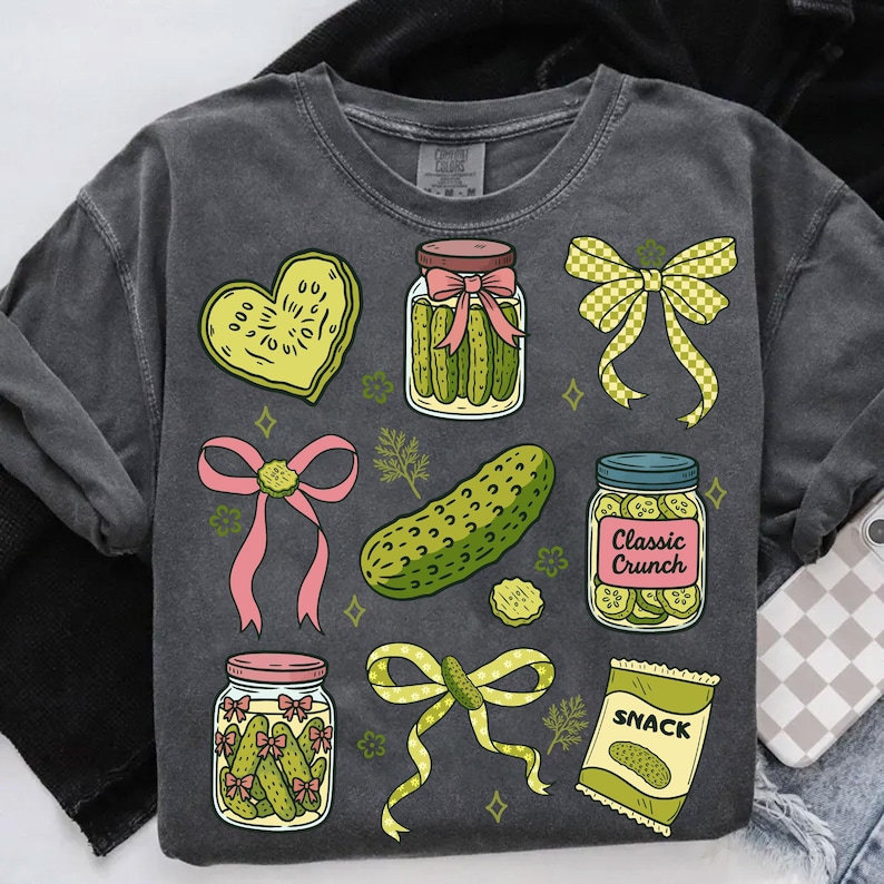 May include: Dark gray sweatshirt featuring a variety of pickle-themed illustrations. The design includes jars of pickles, a pickle-shaped heart, bows, a whole pickle, and a snack bag. The words "Classic Crunch" and "SNACK" are visible.