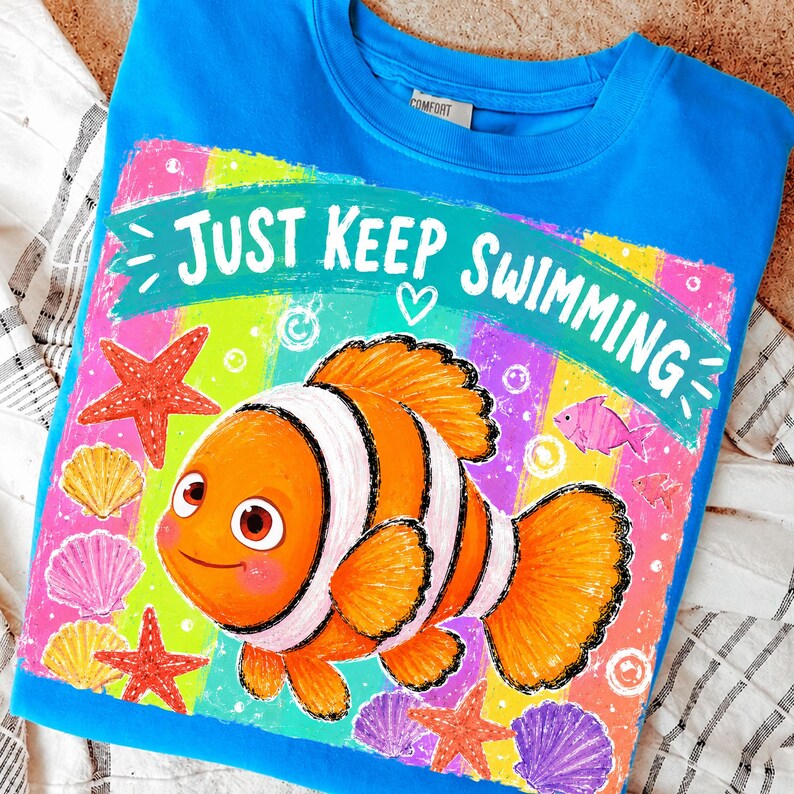 K&ouml;nnte beinhalten: Ein blaues Sweatshirt mit einem farbenfrohen Clownfisch-Motiv, Seesternen, Muscheln und dem Schriftzug "Just Keep Swimming". Der Hintergrund ist ein Regenbogen aus leuchtenden Farben, was ein fr&ouml;hliches Design ergibt.