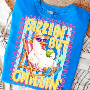 May include: A blue sweatshirt with a graphic of a duck wearing sunglasses and a hat, relaxing in a beach chair while holding a coconut drink. The text "Sizzlin' But Chillin'" is displayed in yellow.
