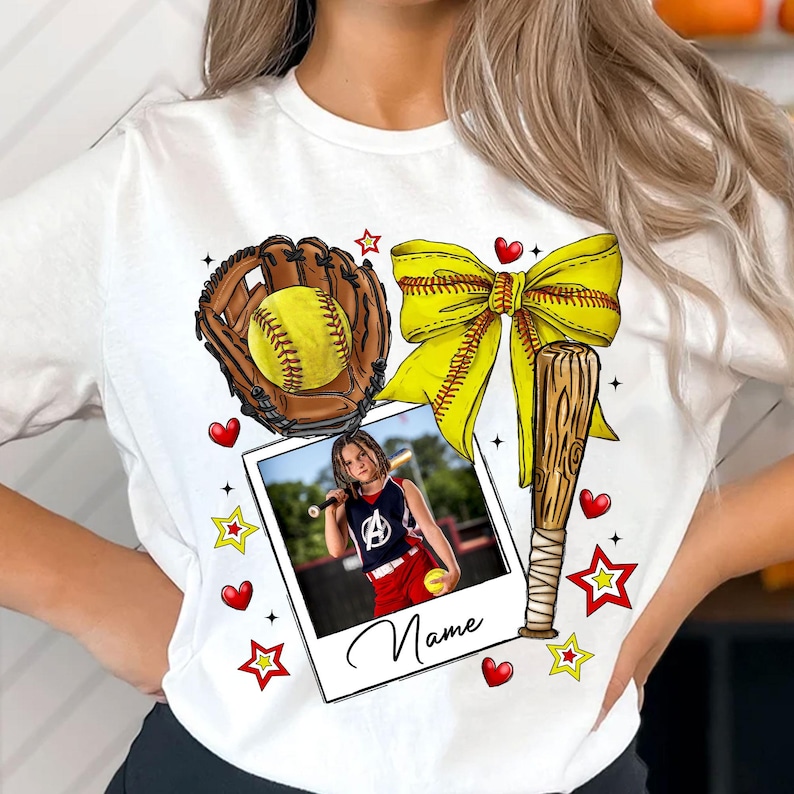 May include: White t-shirt with a softball theme. It features a softball glove, bat, ball, bow, and a photo of a girl playing softball. The text "Name" is at the bottom of the photo.