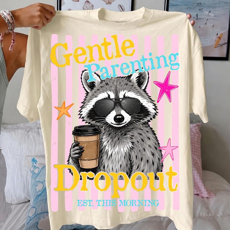 Op de afbeelding: Cr&egrave;mekleurig T-shirt met een afbeelding van een wasbeer met zonnebril en een koffiekopje. Het shirt heeft de tekst "Gentle Parenting Dropout" in geel en blauw, met roze sterren en verticale strepen.