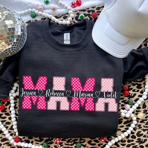 May include: Black sweatshirt with the word "MAMA" in pink and white checkered letters. Below the word "MAMA" are the names Jessica, Rebecca, Mariaa, and Violet. A white trucker hat and disco ball are in the background.