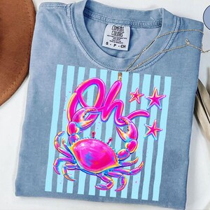 May include: A light blue Comfort Colors t-shirt featuring a vibrant crab graphic with the word "Oh" in a colorful, stylized font. The design includes pink, blue, and yellow accents, with blue vertical stripes and star shapes.