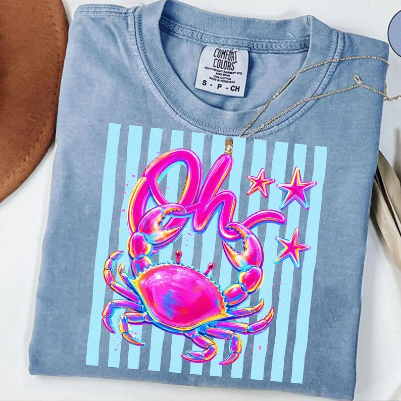 May include: A light blue Comfort Colors t-shirt featuring a vibrant crab graphic with the word "Oh" in a colorful, stylized font. The design includes pink, blue, and yellow accents, with blue vertical stripes and star shapes.