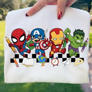 May include: A white sweatshirt featuring a colorful graphic of cartoon superheroes, including Spider-Man, Captain America, Iron Man, and the Hulk, each holding a pencil. Additional school-themed icons like a globe, book, and clock are also present.