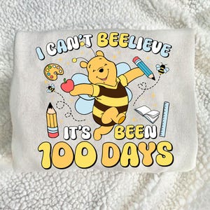 May include: A light gray sweatshirt with a colorful graphic featuring Winnie the Pooh as a bee, holding an apple and pencil. The text reads "I CAN'T BEELEIVE IT'S BEEN 100 DAYS" in yellow and blue.
