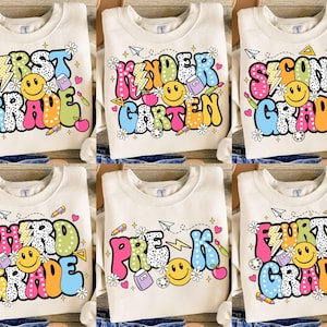 Puede incluir: Sudaderas color crema con textos y gráficos coloridos para diferentes grados: First Grade, Kindergarten, Second Grade, Third Grade, Pre-K y Fourth Grade. Cada diseño incluye una carita sonriente, útiles escolares y flores.