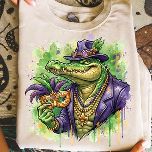 May include: A beige t-shirt featuring a green alligator wearing a purple hat, jacket, and a beaded necklace. The alligator is holding a gold and purple mask. Watercolor paint splatters in green, purple, and yellow are in the background.