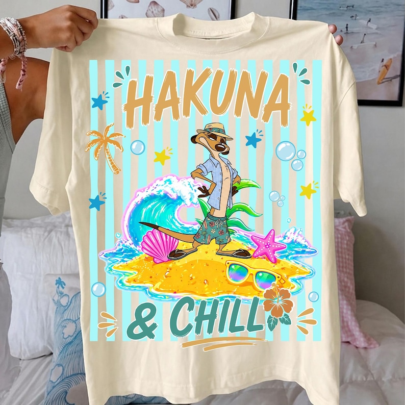 May include: Cream-colored t-shirt featuring a cartoon meerkat on a beach with the words "HAKUNA & CHILL". The design includes a wave, sunglasses, and tropical elements. The shirt has a relaxed fit and short sleeves.