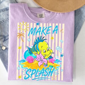 May include: Lilac-colored t-shirt featuring a cartoon fish with the text "MAKE A SPLASH". The design includes a sun, palm tree, and starfish. The shirt is made of a soft material.