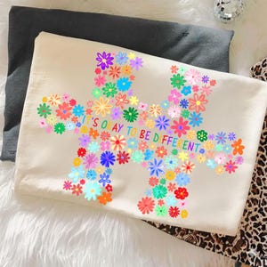 May include: A cream-colored t-shirt featuring a colorful floral design in the shape of a puzzle piece. The text "IT'S OKAY TO BE DIFFERENT" is printed in the center. The shirt is folded on top of a dark gray pillow.