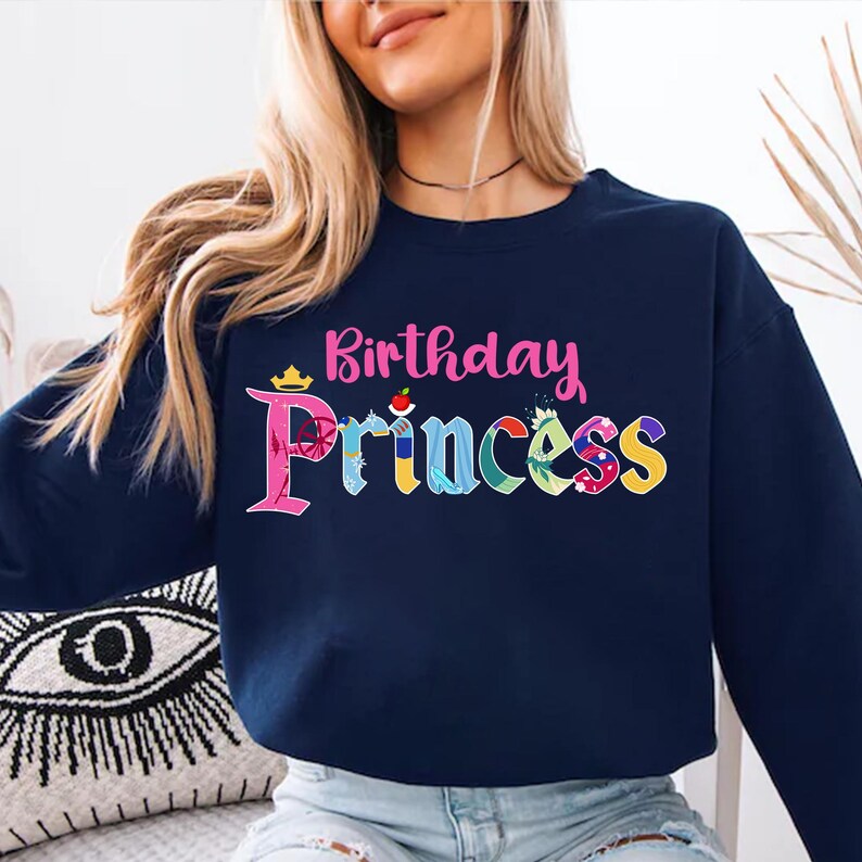 May include: A navy blue sweatshirt with the text "Birthday Princess" in pink and colorful letters. The letters are decorated with images of Disney princesses.
