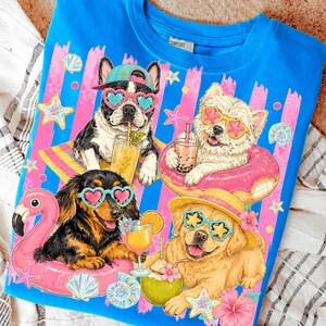 May include: Blue sweatshirt featuring a graphic of four dogs wearing sunglasses and enjoying tropical drinks. The dogs are on inflatable floats with a beach theme, surrounded by starfish and seashells.