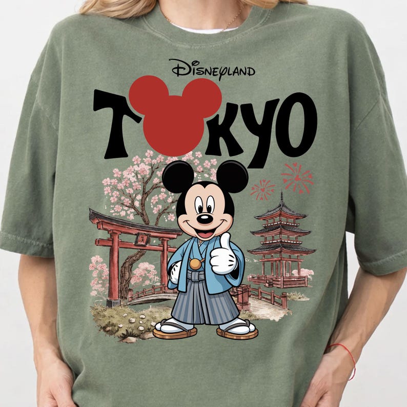 May include: Sage green t-shirt with a graphic featuring Mickey Mouse in traditional Japanese attire, the word "Tokyo" in black, and the Disneyland logo. The design includes Japanese architecture and cherry blossoms.