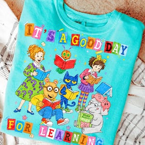 Puede incluir: Una camiseta de color turquesa brillante con un gr&aacute;fico colorido de varios personajes de libros infantiles leyendo. El texto "IT'S A GOOD DAY FOR LEARNING" est&aacute; impreso en letras de bloque multicolores en la parte superior e inferior.