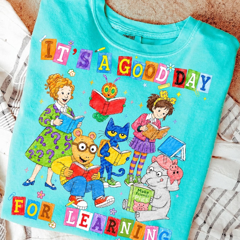 Puede incluir: Una camiseta de color turquesa brillante con un gr&aacute;fico colorido de varios personajes de libros infantiles leyendo. El texto "IT'S A GOOD DAY FOR LEARNING" est&aacute; impreso en letras de bloque multicolores en la parte superior e inferior.