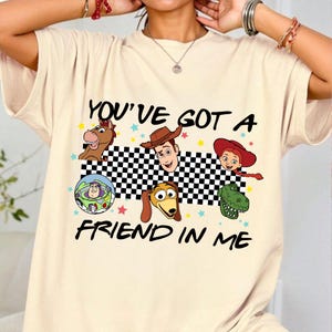 Puede incluir: Camiseta beige con un gr&aacute;fico de personajes animados y el texto "YOU'VE GOT A FRIEND IN ME". El dise&ntilde;o incluye un caballo, un vaquero, una vaquera, un guardabosques espacial, un perro y un dinosaurio.