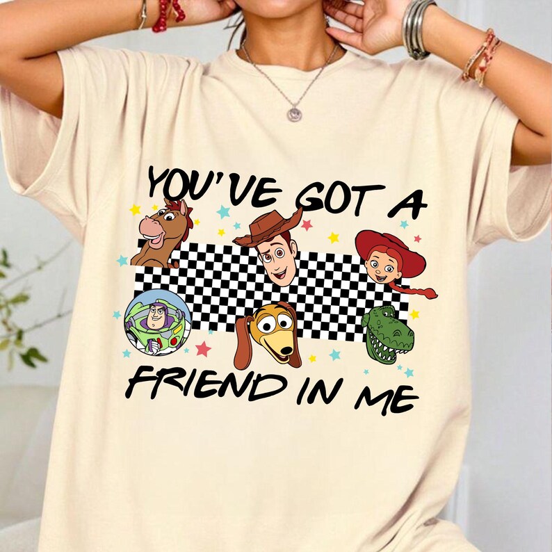 Puede incluir: Camiseta beige con un gr&aacute;fico de personajes animados y el texto "YOU'VE GOT A FRIEND IN ME". El dise&ntilde;o incluye un caballo, un vaquero, una vaquera, un guardabosques espacial, un perro y un dinosaurio.