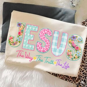 Puede incluir: Camiseta color crema con la palabra "JESUS" en letras bordadas coloridas. Las letras est&aacute;n decoradas con motivos florales, cruces y estampado animal. El texto "The Way The Truth The Life" est&aacute; bordado debajo.