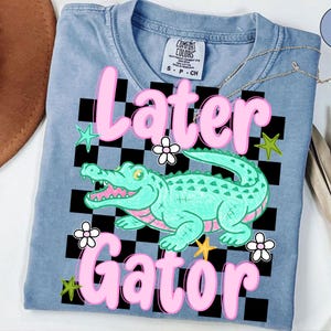 May include: A light blue t-shirt with the words "Later Gator" in pink, featuring a cartoon alligator. The design includes a black and white checkered background, stars, and flowers. The shirt is a Comfort Colors brand.