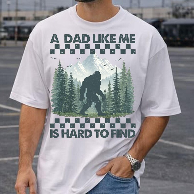 May include: White t-shirt with a graphic of a Bigfoot in a forest scene, with the text "A DAD LIKE ME IS HARD TO FIND". The design includes mountains, trees, and birds, with a checkered border.
