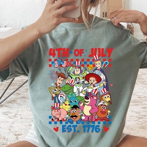 May include: A light blue t-shirt with a graphic of Toy Story characters celebrating the 4th of July. The text "4th of July Est. 1776" is printed on the shirt.