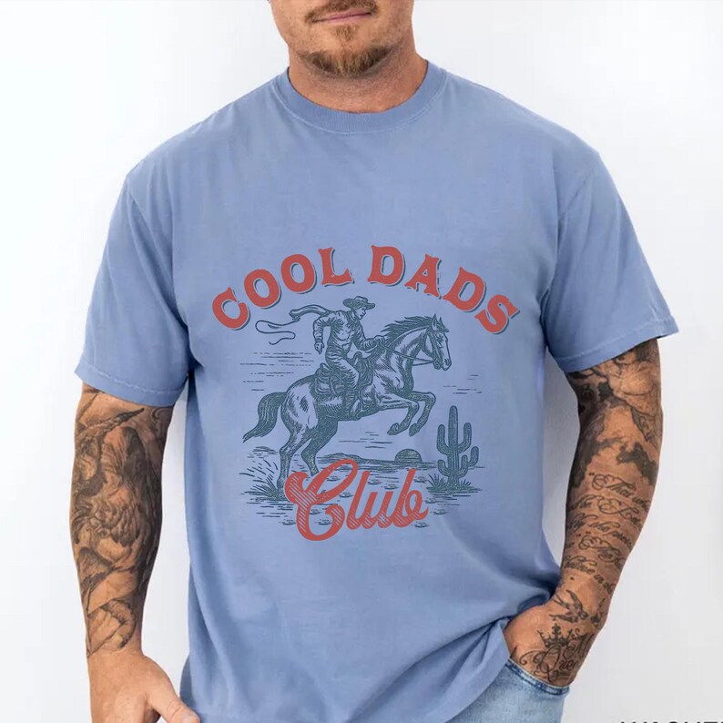 May include: Light blue t-shirt with the text "COOL DADS Club" in red. The graphic depicts a cowboy riding a horse, with a cactus in the background. The shirt is made of a soft material.