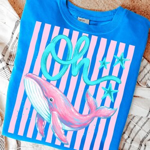 May include: A blue sweatshirt featuring a pink whale illustration with the word "Oh" in teal script. The design includes pink and blue stripes and teal star accents, creating a whimsical and colorful aesthetic.