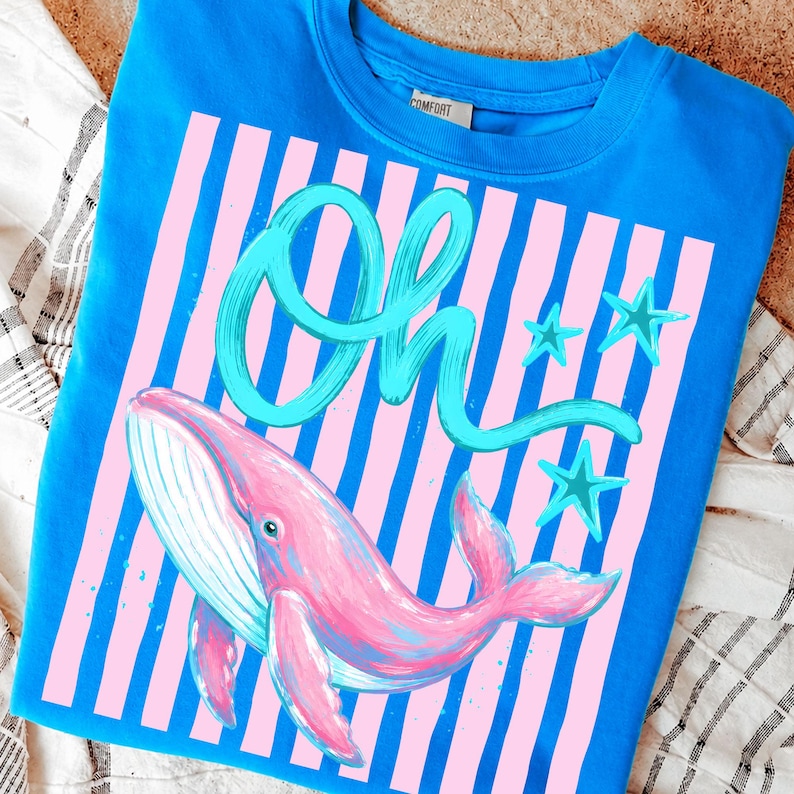 May include: A blue sweatshirt featuring a pink whale illustration with the word "Oh" in teal script. The design includes pink and blue stripes and teal star accents, creating a whimsical and colorful aesthetic.