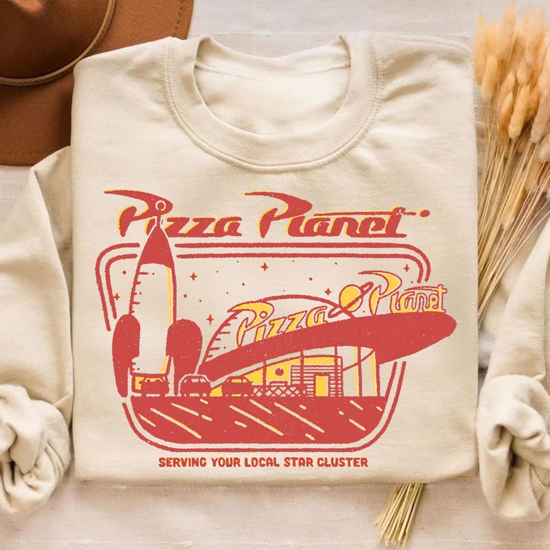 Puede incluir: Sudadera beige con un dise&ntilde;o rojo y amarillo de Pizza Planet, con un cohete y un restaurante. El texto "Pizza Planet" est&aacute; encima del dise&ntilde;o, y "Serving Your Local Star Cluster" debajo.