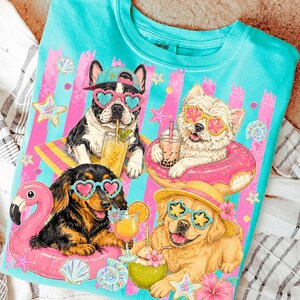 May include: Turquoise t-shirt featuring four dogs enjoying summer. The dogs wear sunglasses and float on inflatable pool toys, with drinks. Pink and blue striped background with seashells and starfish. The word "Comfort" is visible on the collar.