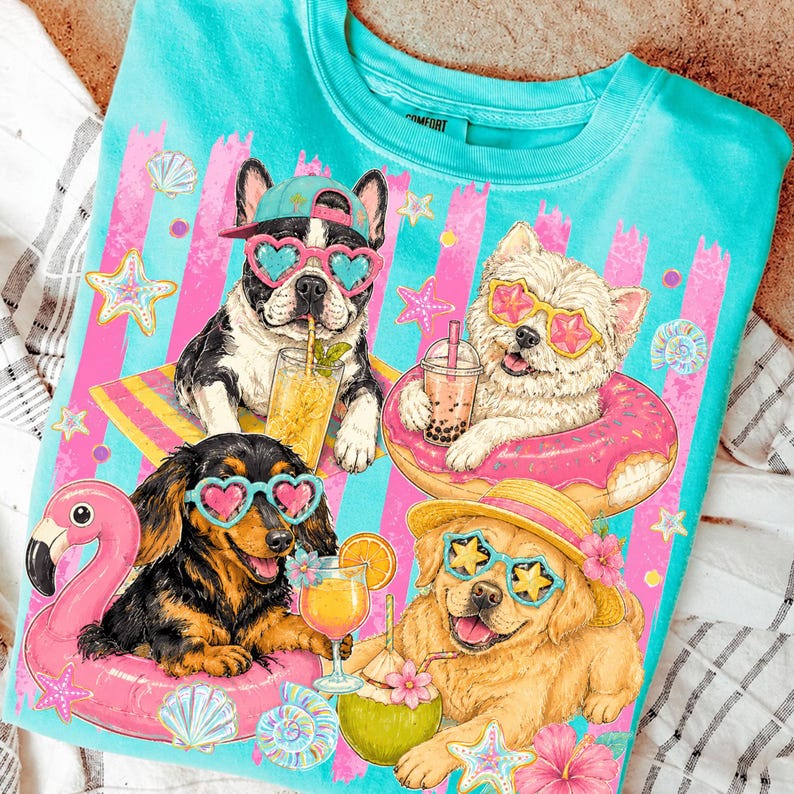 May include: Turquoise t-shirt featuring four dogs enjoying summer. The dogs wear sunglasses and float on inflatable pool toys, with drinks. Pink and blue striped background with seashells and starfish. The word "Comfort" is visible on the collar.