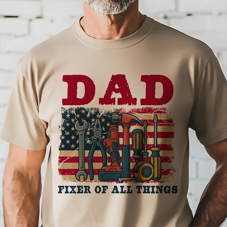 May include: A tan t-shirt with the text "DAD FIXER OF ALL THINGS" in red and black. The text is above a distressed American flag with various tools superimposed on it.