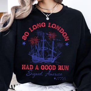 May include: Black sweatshirt with a graphic design featuring a tall ship, fireworks, and text that reads "So Long London, Had a Good Run, Signed, America, 1776". The design is in red and blue.
