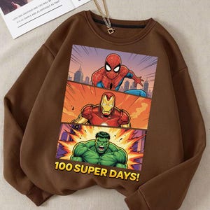 May include: Brown sweatshirt featuring a comic-style graphic of Spider-Man, Iron Man, and the Hulk. The graphic is divided into three panels with a cityscape background. Text at the bottom reads "100 SUPER DAYS!"