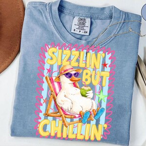 May include: A light blue Comfort Colors t-shirt featuring a cartoon duck wearing sunglasses and a hat, relaxing in a beach chair with a coconut drink. The shirt has the text "Sizzlin' But Chillin'" in a colorful design.