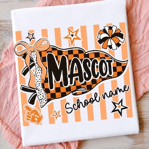 May include: A white t-shirt featuring a graphic print of an orange and black checkered pennant with the text MASCOT and School name. The design includes a bow, a pom-pom, a foam finger, and stars against a background of vertical orange stripes.