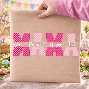 May include: A beige tote bag with the word "MOM" in a pink checkered design. The names Jessica, Rebecca, Marina, and Violet are written below the letters. The bag is held by a person wearing a pink shirt.