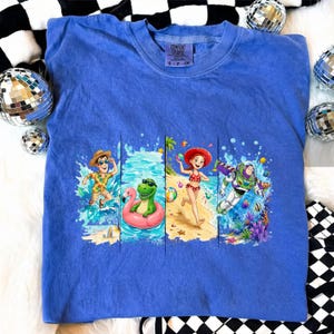 May include: A blue Comfort Colors t-shirt featuring a colorful graphic of animated characters enjoying a beach scene. The graphic includes characters in swimwear, water, and a flamingo float. The shirt has a crew neck and short sleeves.