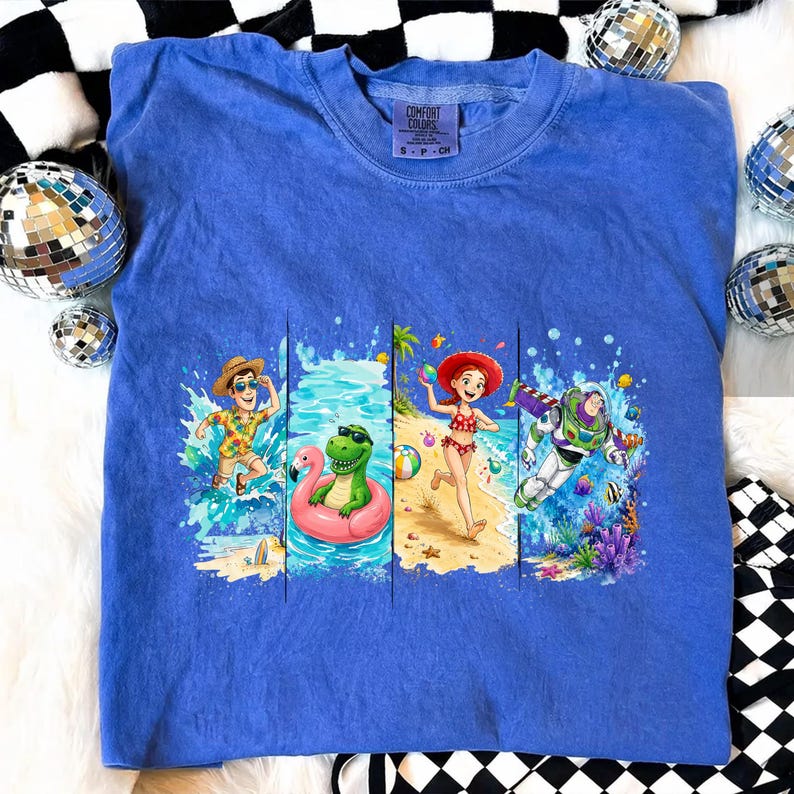 May include: A blue Comfort Colors t-shirt featuring a colorful graphic of animated characters enjoying a beach scene. The graphic includes characters in swimwear, water, and a flamingo float. The shirt has a crew neck and short sleeves.