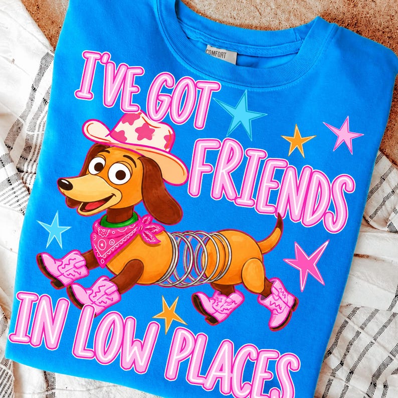 May include: A bright blue t-shirt featuring a cartoon dachshund with a spring body, wearing a pink cowboy hat, a pink bandana, and pink cowboy boots. The text reads I'VE GOT FRIENDS IN LOW PLACES, surrounded by colorful stars in pink, blue, and yellow.