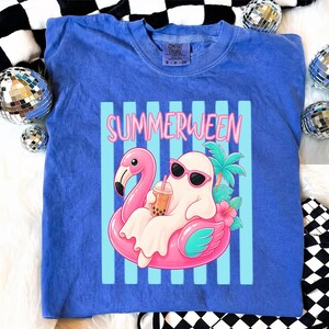 K&ouml;nnte beinhalten: Blaues T-Shirt mit dem Wort "SUMMERWEEN" in Rosa. Das Design zeigt einen Cartoon-Geist mit Sonnenbrille auf einem rosa Flamingo-Schwimmer, der ein Getr&auml;nk h&auml;lt. Das Design umfasst Palmen und Blumen.