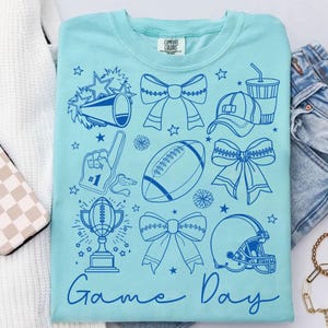 May include: A light blue Comfort Colors sweatshirt with a blue graphic design. The design includes a megaphone, bows, a football, a foam finger, a trophy, a hat, a helmet, and the words "Game Day".