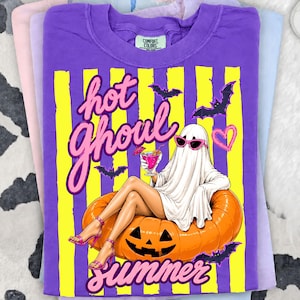 May include: A purple t-shirt featuring a ghost in sunglasses, lounging on a pumpkin and holding a cocktail. The text reads "hot ghoul summer" in pink. The design includes yellow stripes and bat illustrations.