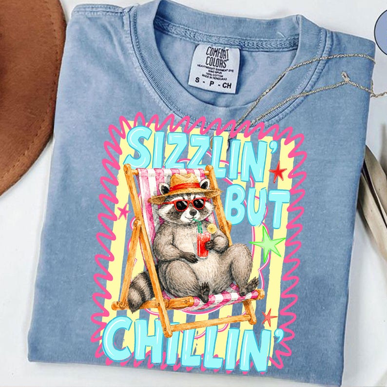 May include: A light blue Comfort Colors t-shirt featuring a raccoon wearing sunglasses and a hat, relaxing in a beach chair with a drink. The shirt has the text "Sizzlin' But Chillin'" in a colorful design.